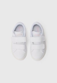 Bílé tenisky se hladkým syntetickým svrškem, vybavené dvěma suchými zipy, měkkou stélkou a minimalistickým designem. Logo Lacoste v růžové barvě.
