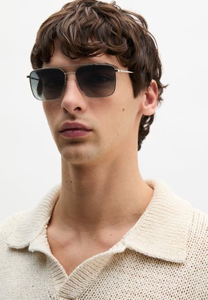 Jonge man met kort bruin haar, draagt donkere rechthoekige zonnebril en een crèmekleurige gebreide trui met kraag, tegen een witte achtergrond.