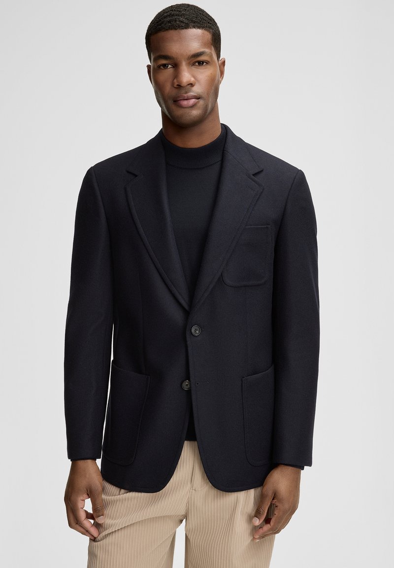 Granatowa marynarka ze strukturalnej tkaniny, z dwoma przednimi kieszeniami, kołnierzem z klapami i dwoma guzikami. Nosi się ją na czarnym golfie.