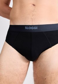 Mann in schwarzen Unterhosen mit einem grauen Bund, auf dem der Markenname "Sloggi" steht, sichtbar bis zur Taille und den oberen Oberschenkeln.