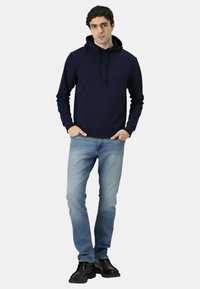 Felpa con cappuccio blu navy con coulisse e fronte testurizzato, abbinata a jeans azzurri e stivali neri, in piedi contro uno sfondo semplice.