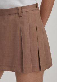 Bruine geplooide rok van een gestructureerde stof, met een platte tailleband en een rits aan de zijkant. Valt boven de knie.