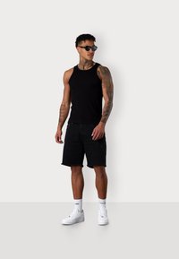 Schwarzes Tanktop und schwarze Denim-Shorts mit fransigem Saum. Accessoires umfassen schwarze Sonnenbrillen und weiße Sneaker mit Logodetails.