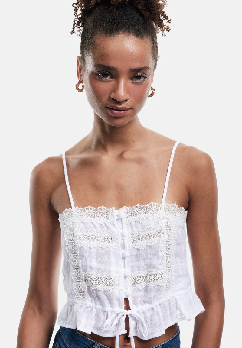 Weißes Cropped Top mit einer Spitzenborte am Halsauschnitt, durchgehenden Knöpfen und einem Rüschensaum. Hergestellt aus einem leichten, strukturierten Stoff. Verstellbare Träger.