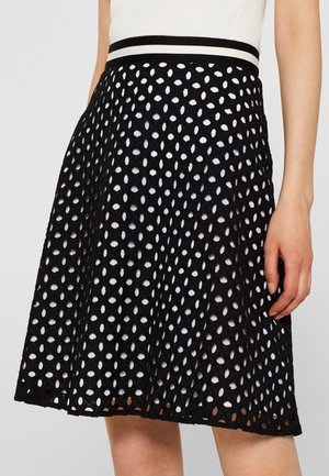 Schwarzer A-Linien-Rock mit weißem, gepunktetem Ausschnittmuster, getragen über einem weißen Unterrock, kombiniert mit einem weißen Oberteil mit schwarz gestreiftem Taillenbund.