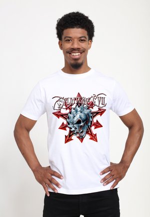 Smilende mand med kort krøllet hår står med hænderne på hofterne iført en hvid T-shirt med rødt stjernemotiv og blå kraniedesign samt teksten "Splinters of Evil".