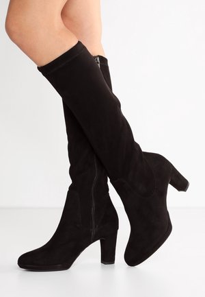Bottes en daim noires montant jusqu'aux genoux, à talons carrés et fermetures éclair latérales, portées sur des jambes croisées sur un fond blanc.