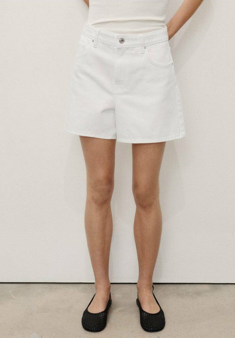 En person står klädd i en figurnära vit topp och vita denimshorts, i kombination med svarta vävda slip-on skor mot en neutral bakgrund.