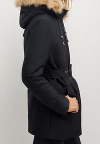 Parka noire à capuche avec bordure en fausse fourrure, taille ceinturée et poches latérales. Présente une texture lisse et des accents métalliques sur les cordons de serrage.