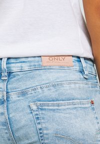 Primer plano de un bolsillo y cinturilla de unos pantalones vaqueros de denim azul claro con una etiqueta beige de la marca ONLY, usados con una camisa blanca metida por dentro.
