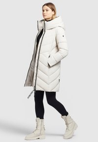 Beige pufferjacka med hög krage, chevronquiltat mönster, sidofickor och svart dragkedja; bärs över svarta leggings och beiga stövlar.