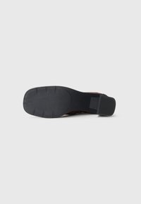 Chaussure à talon en cuir suédé marron avec une semelle extérieure en caoutchouc noir lisse. Présente un bout arrondi et un talon bas en bloc. Détails texturés sur la tige.