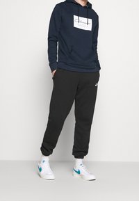 Felpa blu navy con un'etichetta logo bianca, abbinata a pantaloni jogger neri. Sneakers bianche con dettagli blu completano il look.