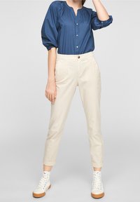 Mujer vestida con pantalones beige de estampado arremangados, blusa azul abotonada con mangas abullonadas y zapatillas altas blancas de pie sobre un fondo gris.
