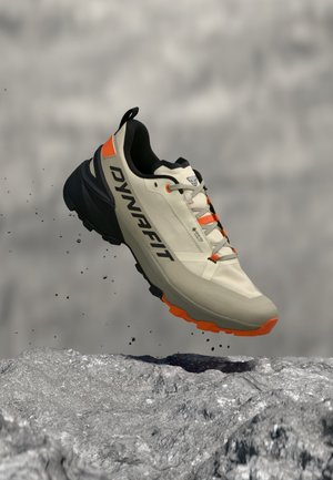 Chaussure de trail Dynafit beige clair et orange en plein vol au-dessus d’un sol rocheux avec des particules de poussière.