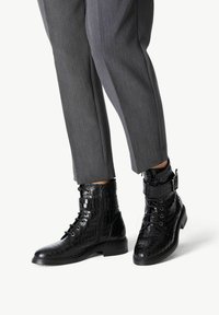 Bottes noires en cuir à la cheville avec un motif crocodile, présentant des lacets, une fermeture éclair sur le côté et une boucle décorative. Portées avec un pantalon gris ajusté.