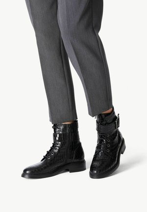 Bottes noires en cuir à la cheville avec un motif crocodile, présentant des lacets, une fermeture éclair sur le côté et une boucle décorative. Portées avec un pantalon gris ajusté.