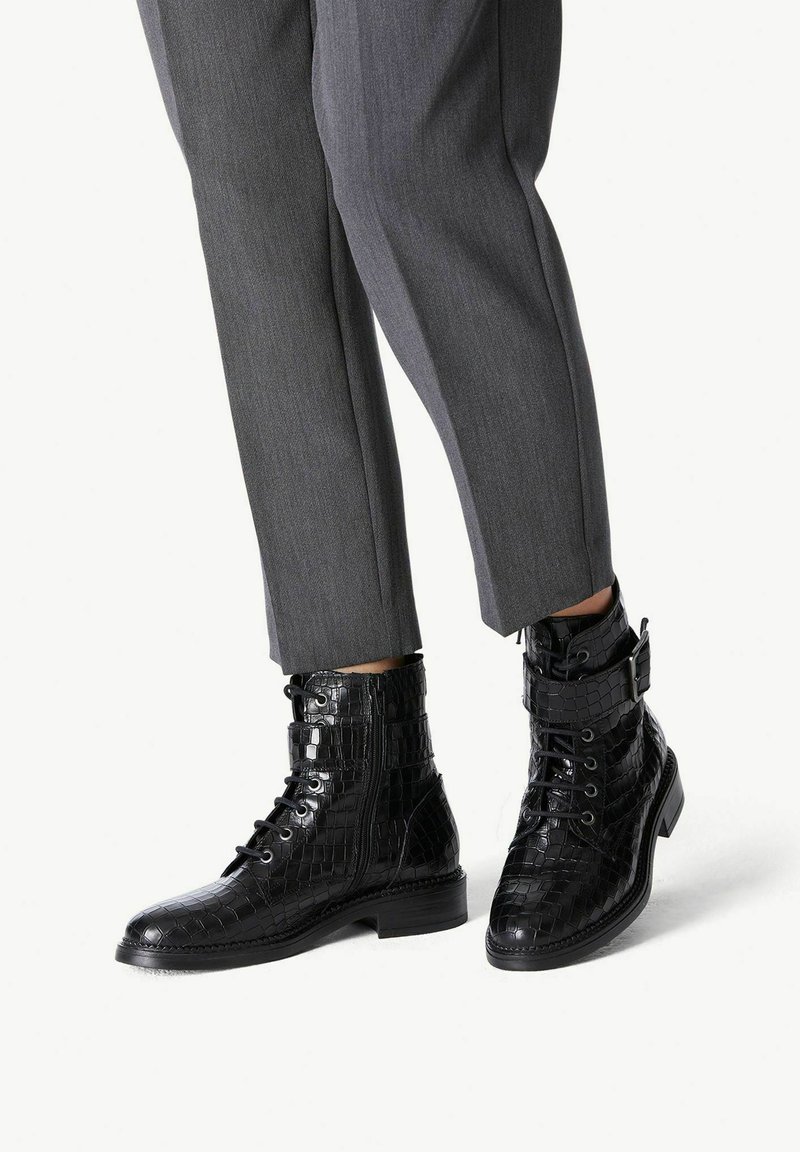 Bottes noires en cuir à la cheville avec un motif crocodile, présentant des lacets, une fermeture éclair sur le côté et une boucle décorative. Portées avec un pantalon gris ajusté.