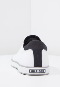 Sneaker bianca in tela con tallone nero e etichetta "HILFIGER" su una suola bianca, vista dal retro su una superficie bianca.