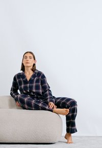 Žena v tmavých károvaných pyžamách sedí naboso na svetlej textúrovanej sedačke proti jednoduchej bielej stene.
