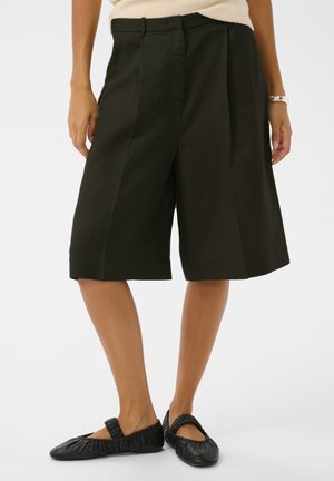 Femme portant un short vert foncé mi-cuisses, un haut beige, des ballerines noires style Mary Jane et une montre-bracelet argentée, debout devant un fond blanc.