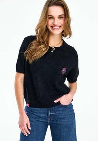 Nasmejana žena s dugom svijetlosmeđom kosom koja nosi crnu majicu kratkih rukava s cvjetnim aplikacijama i plave traperice, stoji s jednom rukom u džepu.