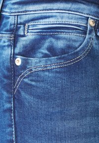 Detailní záběr modrých džínových kalhot s bílým prošíváním a kovovým nýtem blízko okraje kapsy.