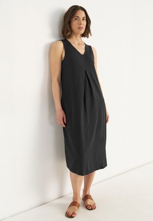 Schwarzes ärmelloses Kleid mit V-Ausschnitt, das über ein plissiertes Design und eine lockere Passform verfügt, aus leichtem Stoff und knielang. Dazu werden braune Rutschsandalen getragen.