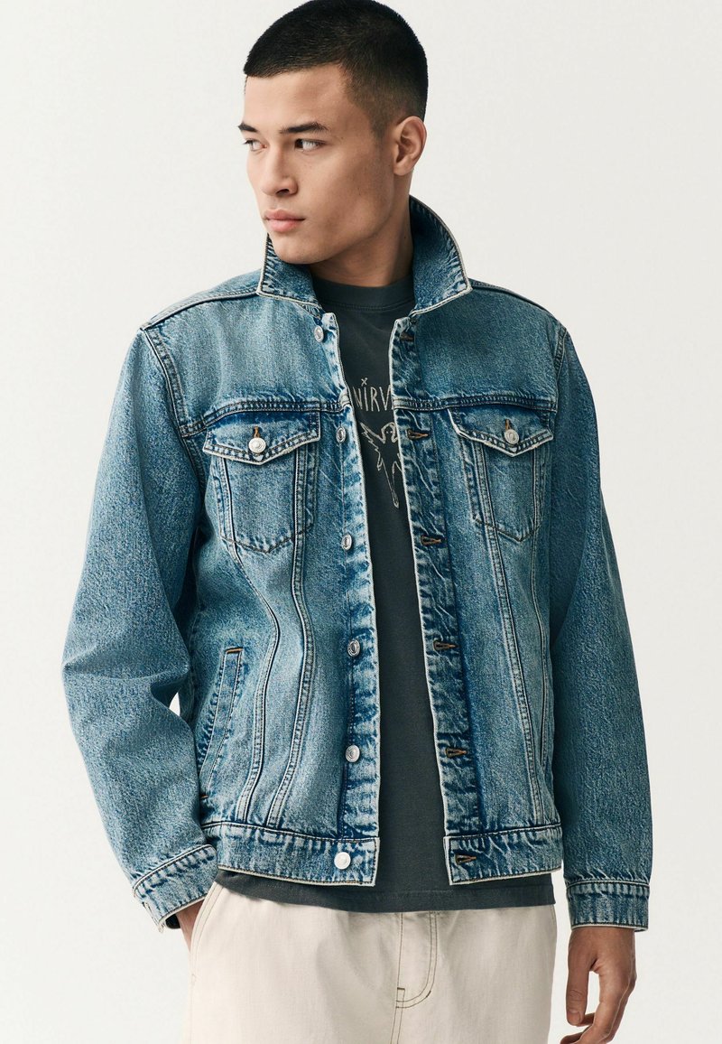 Denimjakke i lyseblå med to forlommer og metalknapper. Har en klassisk krave og en let falmet struktur.
