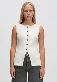Cremefarbener, ärmelloser Oberteil mit rundem Halsausschnitt, ausgestattet mit einer sechs Knöpfe langen Knopfleiste auf der Vorderseite und einem gespaltenen Saumdesign. Weicher, strukturierter Stoff.