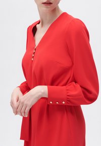 Rote Bluse mit V-Ausschnitt, goldenen Knopfakzenten, langen Puffärmeln und einem seitlichen Bindedetail. Glattes Material mit einer Tasche auf der linken Seite.