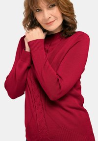 Frau mit hellbraunen, lockigen Haaren, die einen roten, grob gestrickten Pullover trägt, hält ihre Hände nahe ihrem Gesicht vor einem schlichten Hintergrund.