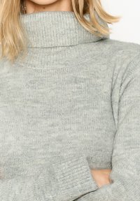 Svetra s svetlo sivo turtleneck ovratnikom iz teksturiranega pletenega materiala, ki ima visok ovratnik in oprijete rokave z rebrastimi manšetami.