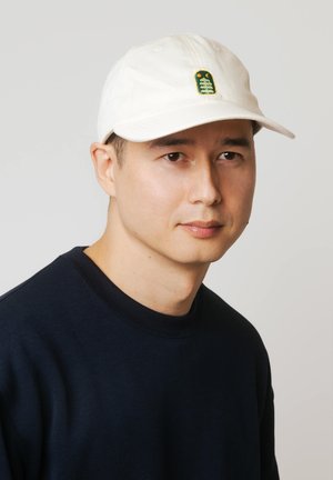 Mężczyzna w białej czapce z małą zielono-pomarańczową naszywką sowy, ubrany w ciemnogranatową koszulę, neutralne jasnoszare tło.