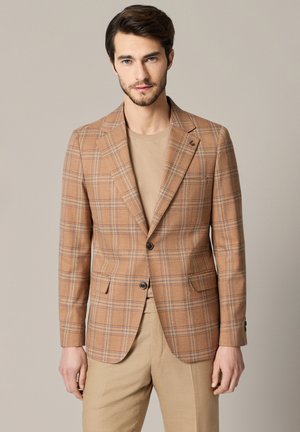 Mężczyzna w beżowej kraciastej marynarce noszonej na beżowej koszuli i spodniach, stojący na tle jednolitego neutralnego tła, patrzy w obiektyw.