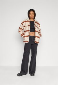 Overdimensjonert fleecejakke i krem og rust med striper og sherpa-krage, kombinert med en svart crop-top og svarte vide bukser samt tresko.