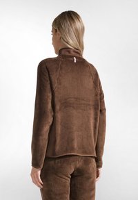 Pullover in pile marrone con colletto alto, superficie testurizzata e una cucitura posteriore sottile. Presenta un piccolo tag sul collo.