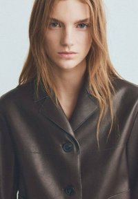 Brązowa skórzana marynarka o strukturalnym kroju, z klapami z wycięciem, czarnymi guzikami i gładką fakturą. Modelka z długimi włosami.