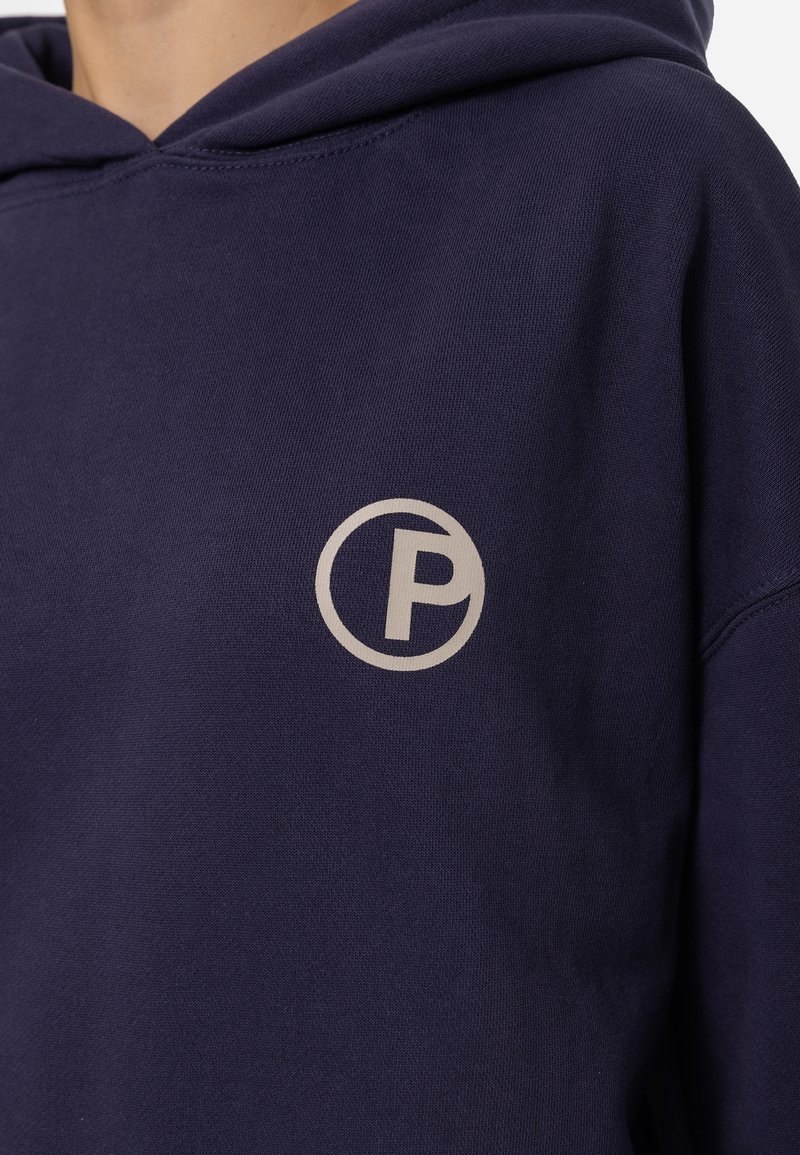 Sweat à capuche bleu marine avec un petit logo blanc en forme de cercle contenant un "P" sur la partie gauche de la poitrine, porté par une personne.