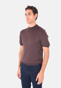 Uomo in maglietta a maniche corte marrone a maglia e pantaloni blu navy, in piedi e che guarda leggermente di lato su uno sfondo bianco uniforme.
