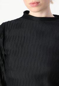 Juodas tekstūruotas viršus su aukštu kaklu ir banguota raštu audinyje, sukuriantis subtilų bangų efektą.