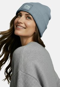 Light blue ribbed knit beanie with a square patch featuring a camera icon. The individual is wearing a grey sweater, partially visible.