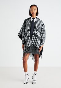 Čierno-biely károvaný pončo s detailmi s třásňami prehodené cez vzorované šaty. Modelka nosí kravatu, ponožky a sandále na vysokom opätku.