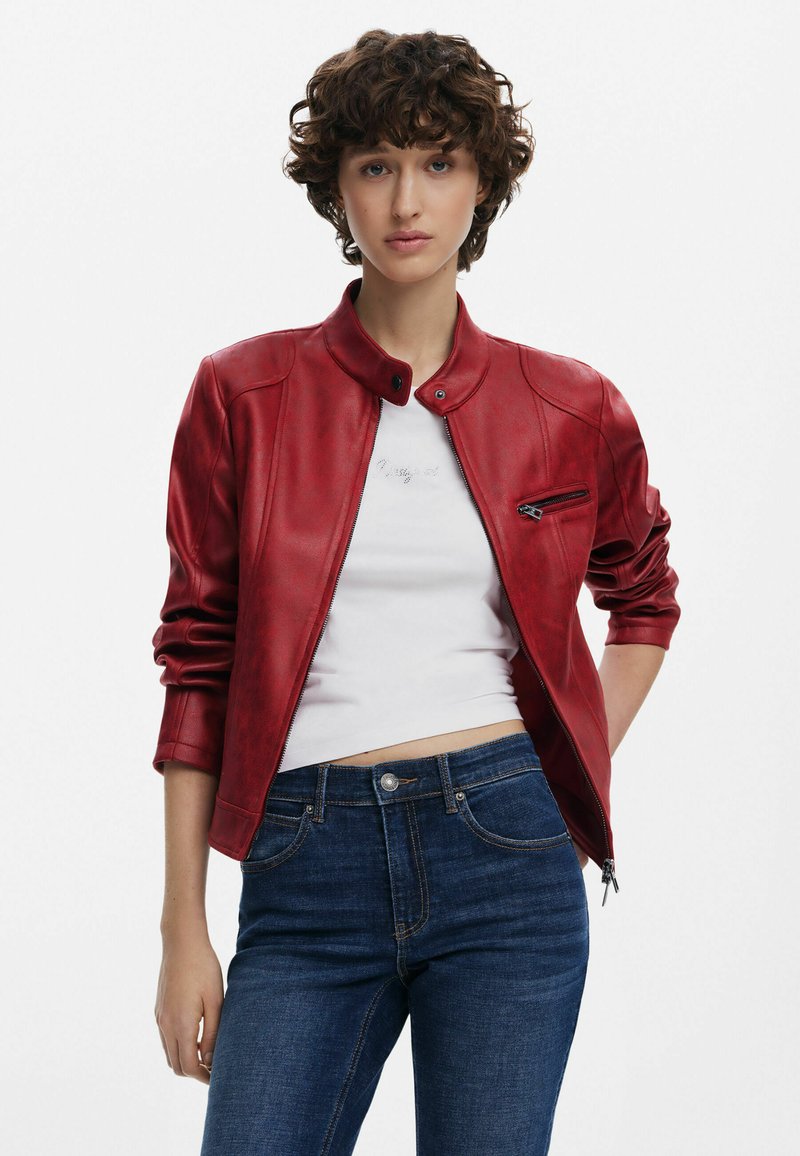 Rote Lederjacke mit Stehkragen, Frontreißverschluss und Reißverschlusstasche auf der Brust. Getragen über einem weißen Hemd mit kurzgeschnittenen Ärmeln und dunkelblauen Jeans.