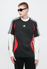 Μαύρο κοντομάνικο t-shirt με κόκκινα στοιχεία, λογότυπο Adidas στο στήθος και λευκές ρίγες στους ώμους. Το μοντέλο φορά μαύρα γυαλιά ηλίου και σκούρο τζιν.