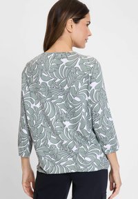 Bluse mit grünem Blättermuster und Dreiviertelärmeln, aus weichem Stoff, mit runder Ausschnitt und weißem Hintergrund.