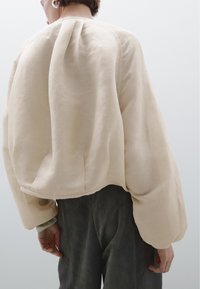 Persoon in een oversized beige linnen jasje met verlaagde schouders en brede mouwen, gecombineerd met donkere wijde broek, tegen een effen achtergrond.