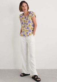 Bluză cu mâneci scurte, cu un model floral în culori galben, mov și verde; asortată cu pantaloni din in albi și sandale negre, având o croială lejeră.