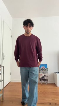 Ung mann med krøllete hår iført en mørkerød langermet skjorte og blå jeans som står på en tregulv i et hvitt rom.
