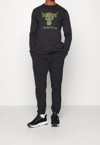 Man med svart långärmad tröja med grön tjurlogo och texten "Project Rock", svarta joggerbyxor, svart-vita sneakers, händerna i fickorna.